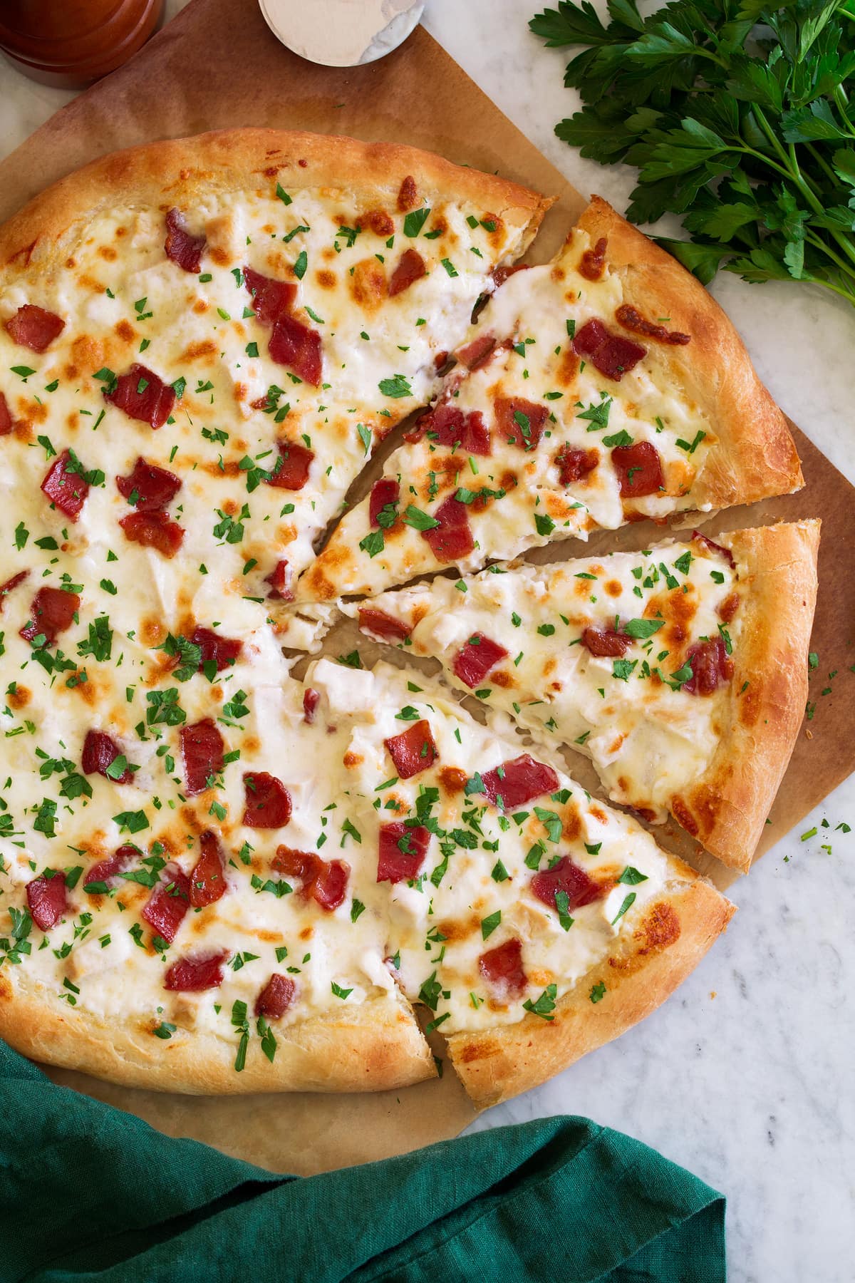 Overhead photo of chicken alfredo pizza cut into slices. Pizza is set over parchment paper and has a green cloth to the side.