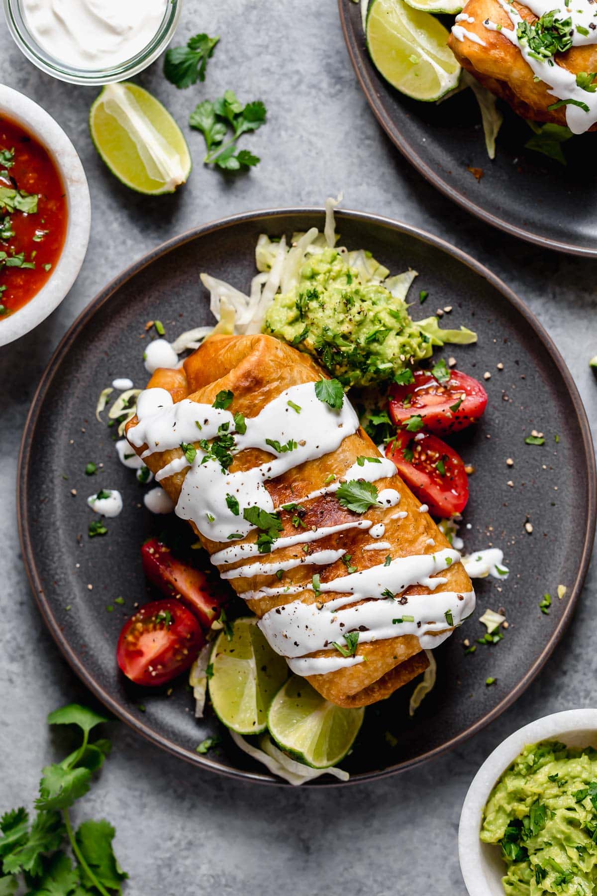 Chicken Chimichanga on a dark plate garnished with guacamole, sour cream, tomatoes and cilantro.