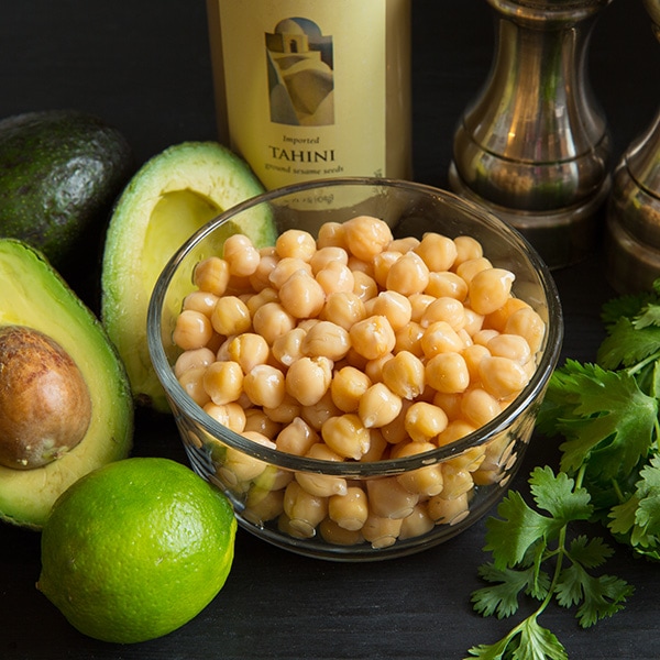 A bowl of chickpeas with other ingredients to make avocado hummus at the side