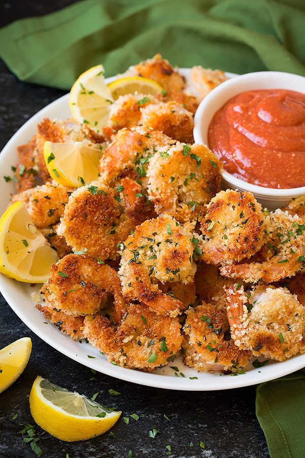 Shrimp with panko breading served on a white plate with a side of cocktail sauce and lemon wedges..