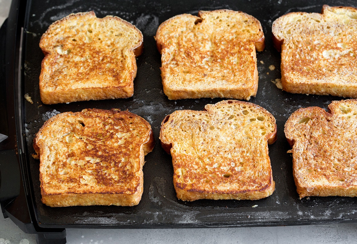 Showing how to cook french toast. Six slices with golden brown tops are being cooked on an electric griddle.