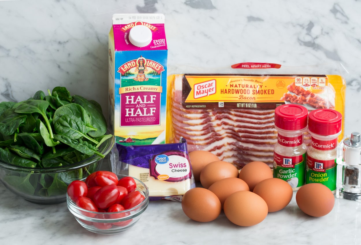 Ingredients needed to make a frittata shown here including eggs, bacon, swiss cheese, spinach, tomatoes, half and half, garlic powder, onion powder, salt and pepper.
