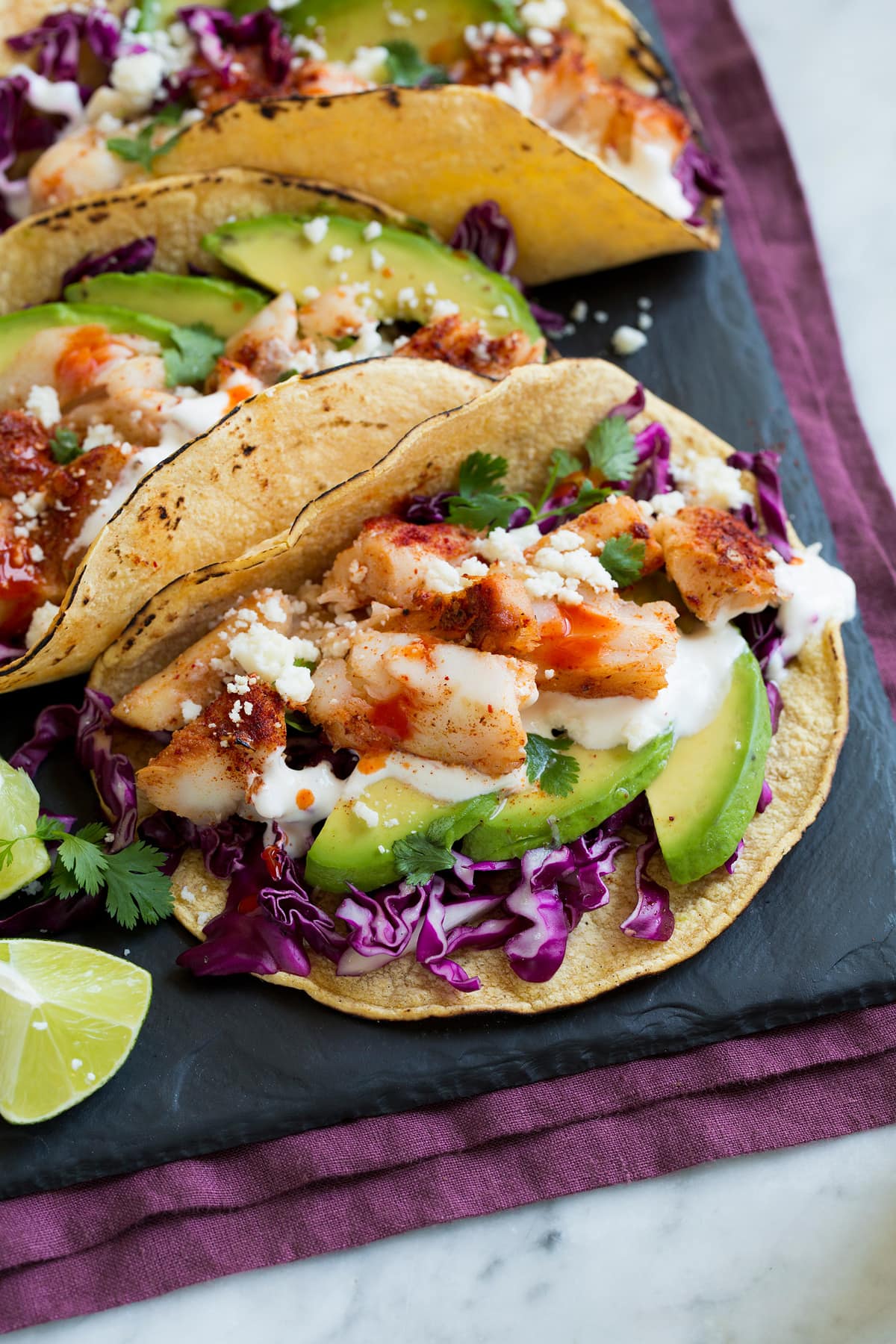 Close up image of fish tacos layered with purple cabbage, avocado, fish taco sauce, cod, talapia, and cotija.