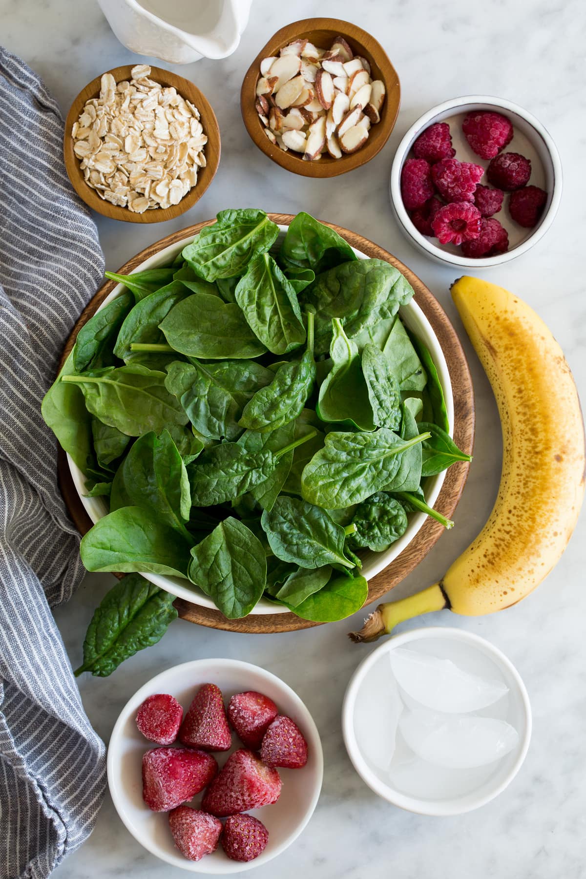 Ingredients needed to make green smoothie shown here including spinach, strawberries, raspberries, bananas, almonds, oats, water, and ice.