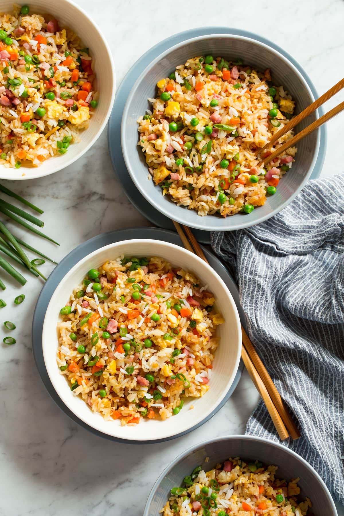 Four servings of fried rice shown overhead in white serving bowls set over blue plates with chopsticks to the side.