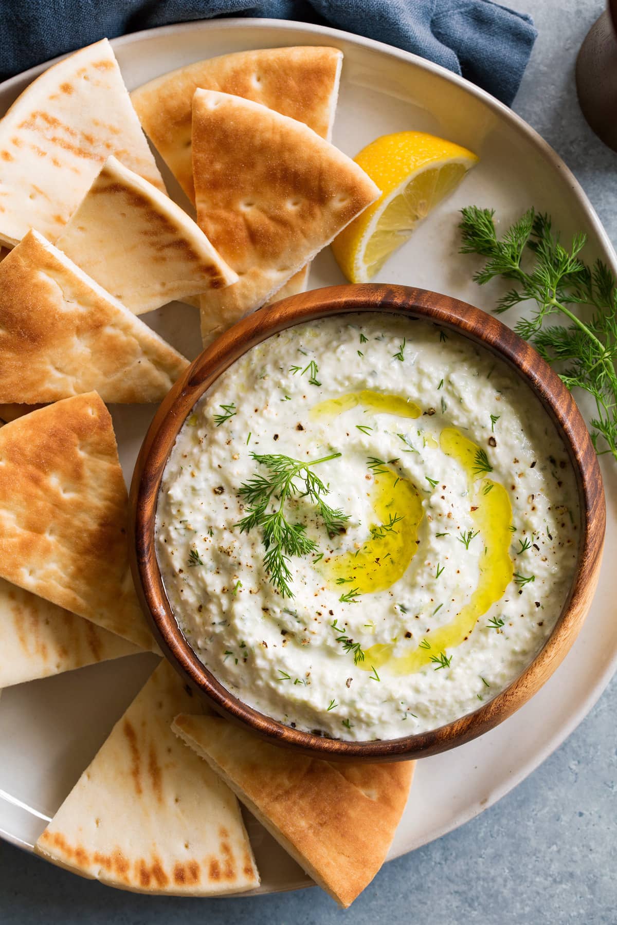 Overhead image of tzatziki sauce in a wooden bowl on a white plate with pita bread for dipping.