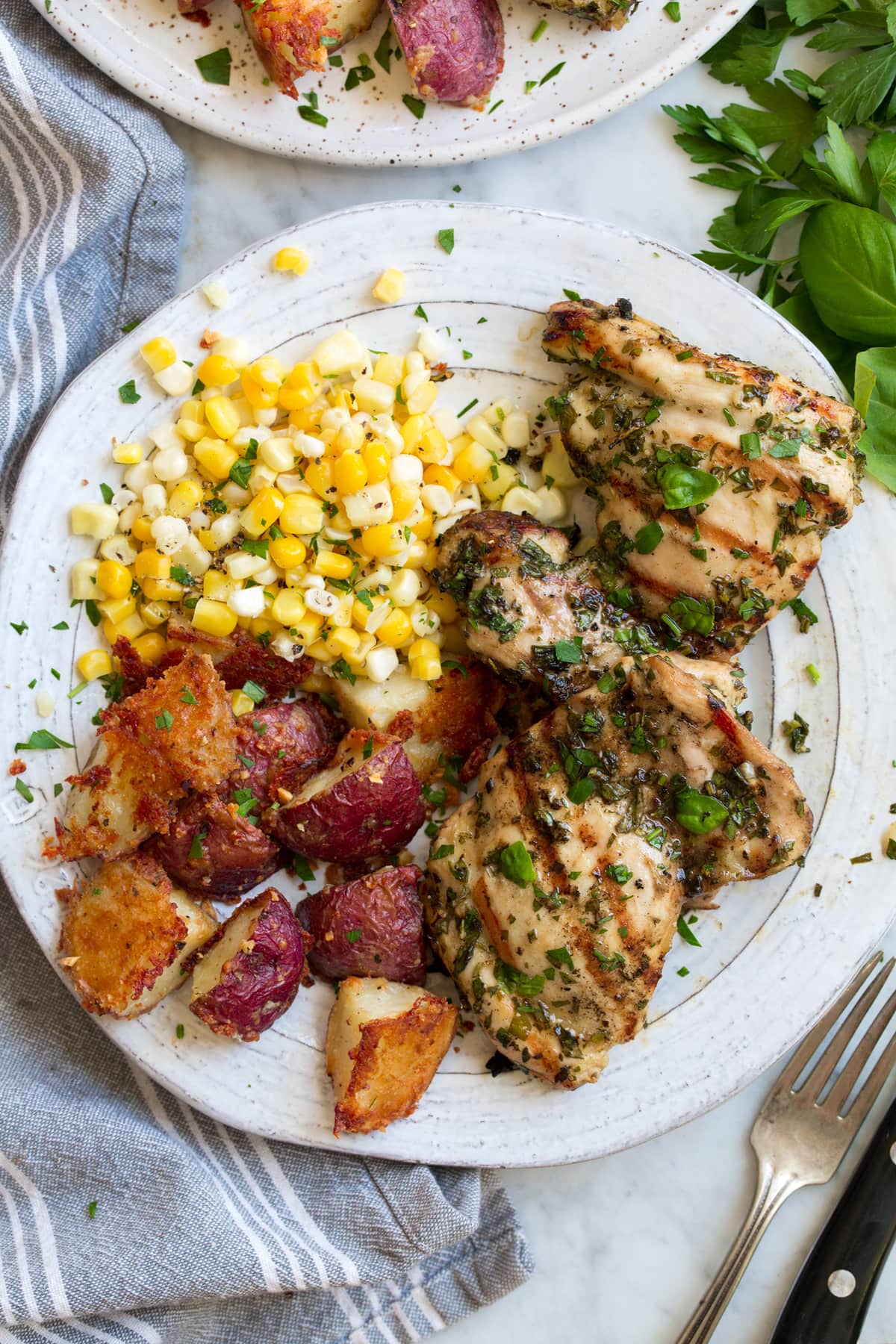 Overhead image of garlic and herb marinated grilled chicken thighs with corn and roasted red potatoes as sides. Food is resting on a white plate set over a marble surface.