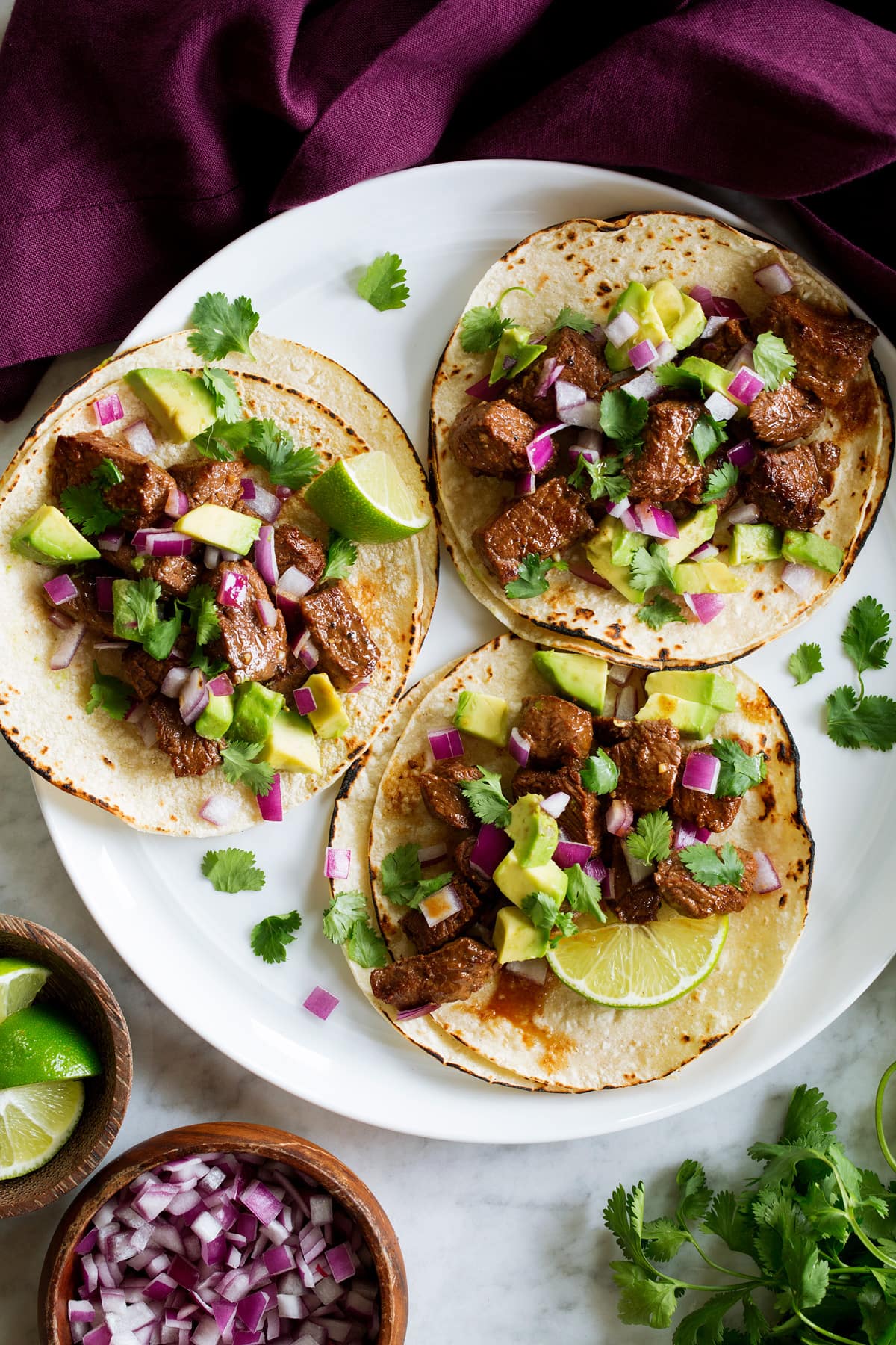 Three steak tacos on a white serving plate with a purple cloth to the side.
